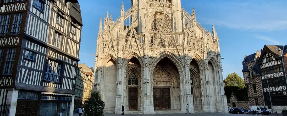 The Gothic-style Saint-Maclou Church in Rouen, featuring a grand entrance with detailed stone carvings, flanked by half-timbered medieval buildings.