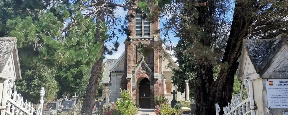 The entrance of Saint-Martin Church framed by trees and an iron gate, with a mix of stone and brick architecture and a well-maintained garden.