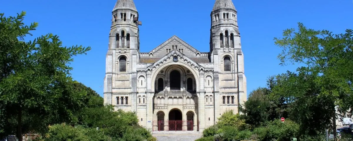 A front view of Saint-Martin Church in Périgueux, characterized by its two tall towers and Romanesque architectural style. The building is framed by greenery, with a clear blue sky overhead.