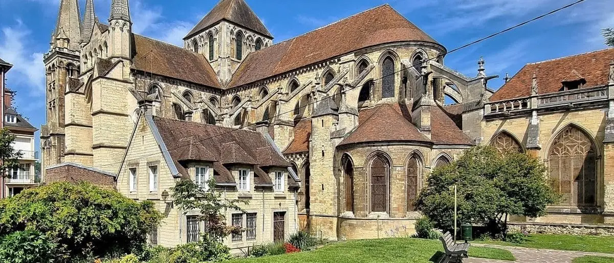 The Saint Pierre Cathedral in Lisieux, displaying its impressive Gothic architecture with intricate stonework, flying buttresses, and pointed arches. The structure is complemented by a well-kept lawn and a few surrounding buildings.