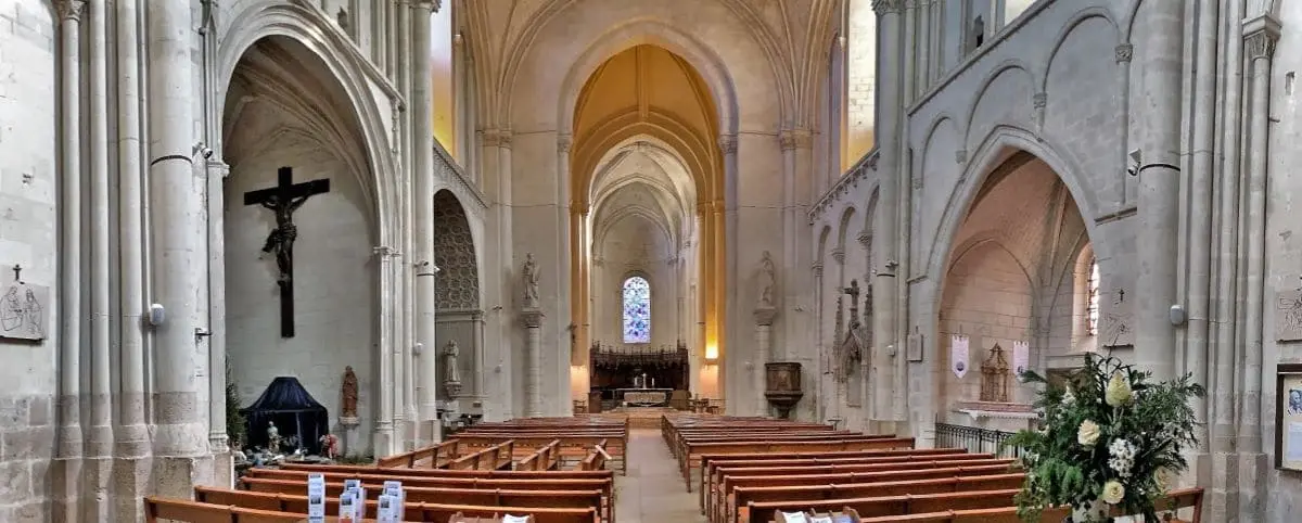 The nave of Saint Pierre Church in Saumur, featuring tall arches, wooden pews, a large crucifix, and a bright stained glass window at the far end.