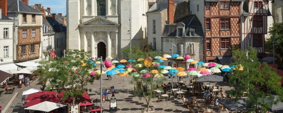 Vibrant scene of Saumur town center with colorful umbrellas hanging overhead, outdoor café tables, and historic buildings lining the square.