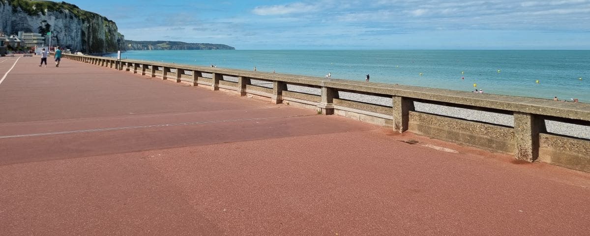 The seafront promenade in Dieppe, featuring a wide paved walkway parallel to the beach, with cliffs visible in the background and a clear blue sky.
