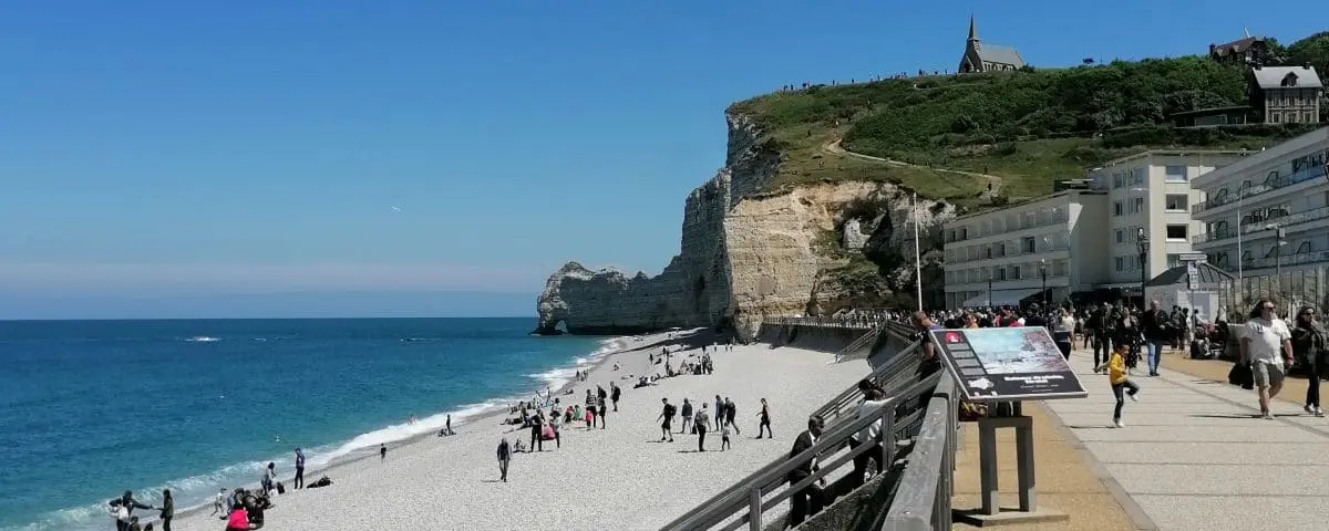 Busy seafront in Etretat with people strolling along the promenade, white cliffs, and the sparkling sea under a bright blue sky.