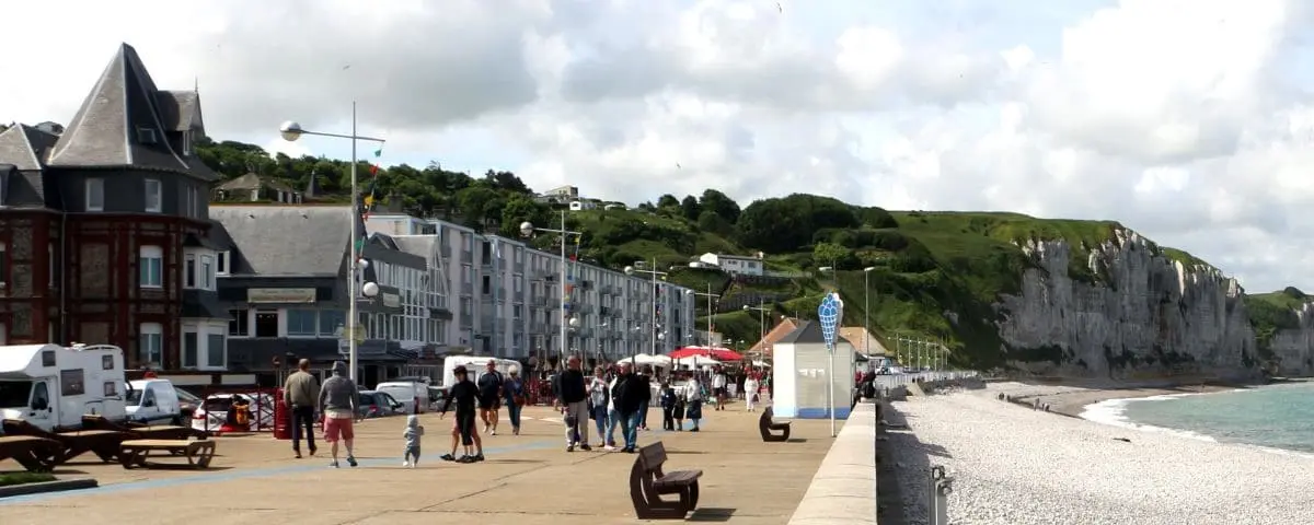The seafront promenade in Fécamp, France, with people walking along the shore, charming seaside buildings, and the white cliffs in the distance.