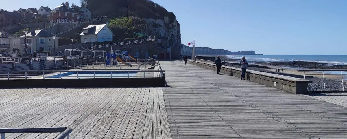 A wooden boardwalk along the seafront with a few people strolling. The scene includes a view of cliffs and the ocean under a clear sky.