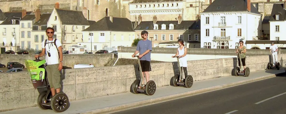 A group of tourists riding Segways on a sunny day, lined up along a stone bridge. Historic buildings and the town of Amboise can be seen in the background.