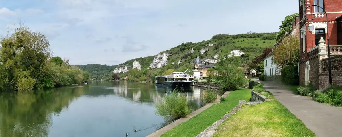 A serene scene of the Seine River as it winds through the lush greenery and hills of Les Andelys, with boats docked along the riverbank and quaint houses in the background.