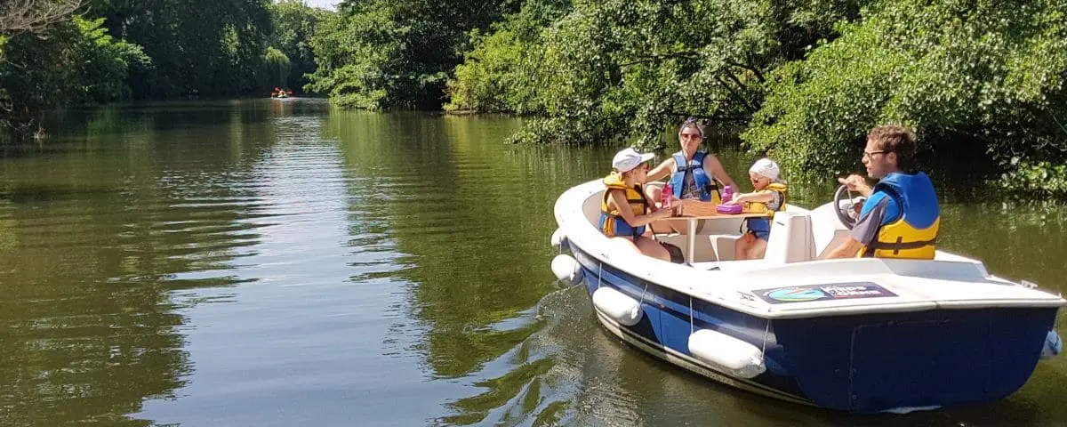 A family enjoying a ride in a self-drive river boat on a calm, tree-lined river in Caen. Everyone is wearing life jackets, and the children are happily engaged in conversation.