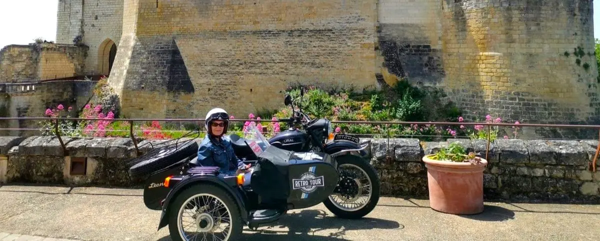 Person wearing a helmet and sunglasses, sitting on a retro sidecar motorcycle parked near a historic stone wall, with blooming flowers and greenery in the background.