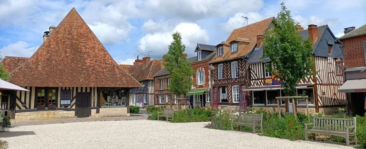 A picturesque square in Beuvron-en-Auge, featuring charming timber-framed buildings, a stone building with a steeply pitched roof, and a well-manicured garden area with benches.