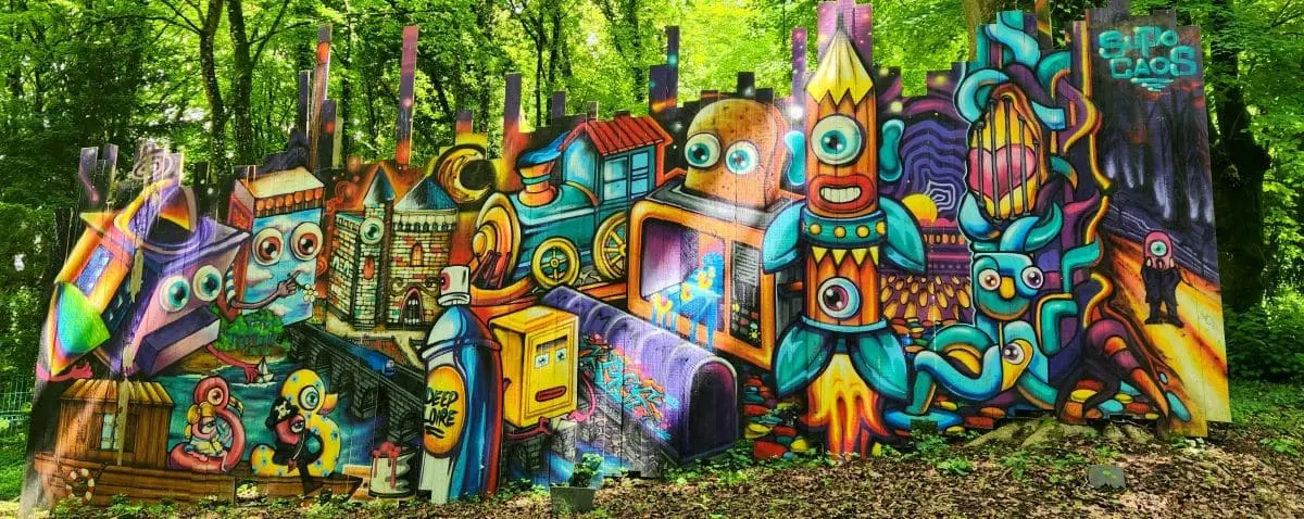 Vibrant and colorful street art mural featuring whimsical characters and imaginative scenes, set in a lush, green forested area.