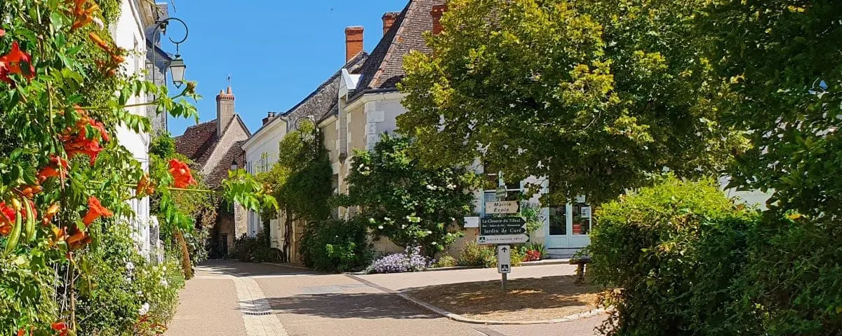 A charming street in Chédigny lined with white stone houses adorned with vibrant flowers and greenery, leading to a signpost pointing to local attractions.