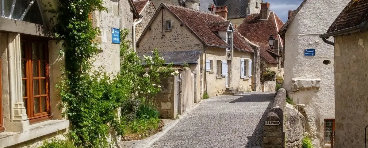 A charming cobblestone street in Montrésor lined with historic stone houses covered in greenery and flowers, leading towards the village center.