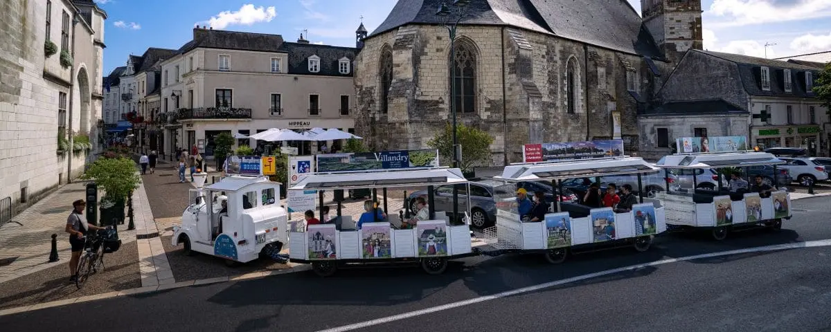 A small tourist train with open-air carriages, filled with visitors, parked on a street near historic buildings and a church. The train is used for guided tours around the town.