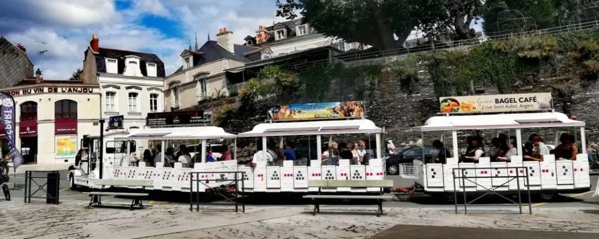 White tourist train with open carriages filled with passengers, parked in a historic part of Angers, near quaint buildings and cafes.