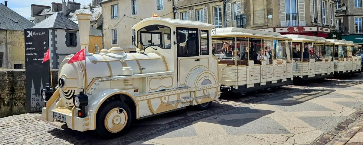 A charming white and gold tourist train with passengers on board, parked on a cobblestone street in Bayeux with buildings in the background.