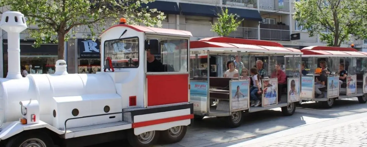 A white and red tourist train with passengers seated in open carriages, traveling through a street in Caen. The surrounding shops and trees indicate a lively urban setting.