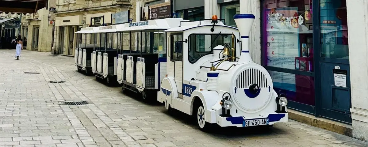 A white and blue tourist train parked on a cobbled street in Périgueux. The train is designed for sightseeing, with carriages featuring open sides for better views of the town's attractions.