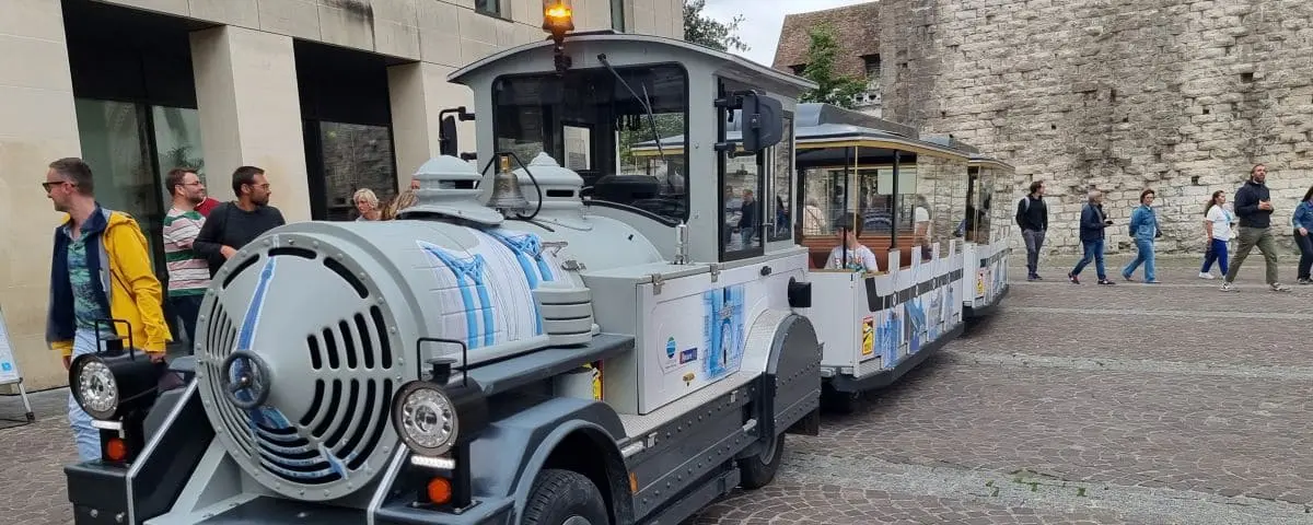 A small, tourist train in Rouen, designed to resemble a vintage locomotive, with open carriages for passengers, parked near a historical stone wall with tourists nearby.
