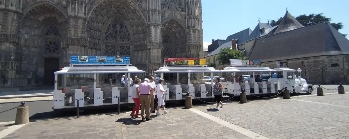 A tourist train with open carriages parked in front of a historic building in Tours, France, with passengers boarding and alighting under a sunny sky.