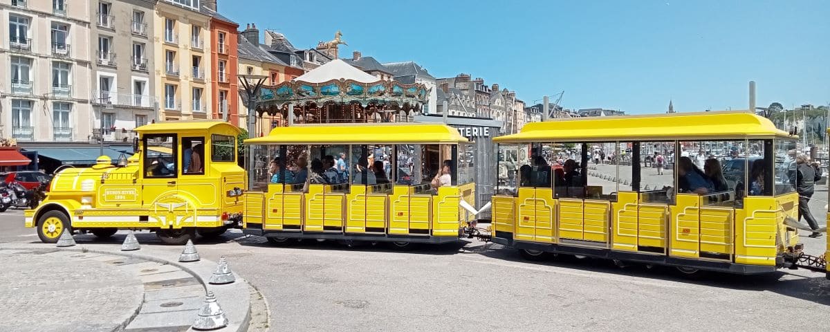 A bright yellow touristic train in Dieppe, with passengers onboard, driving past colorful buildings and a vintage carousel on a sunny day.