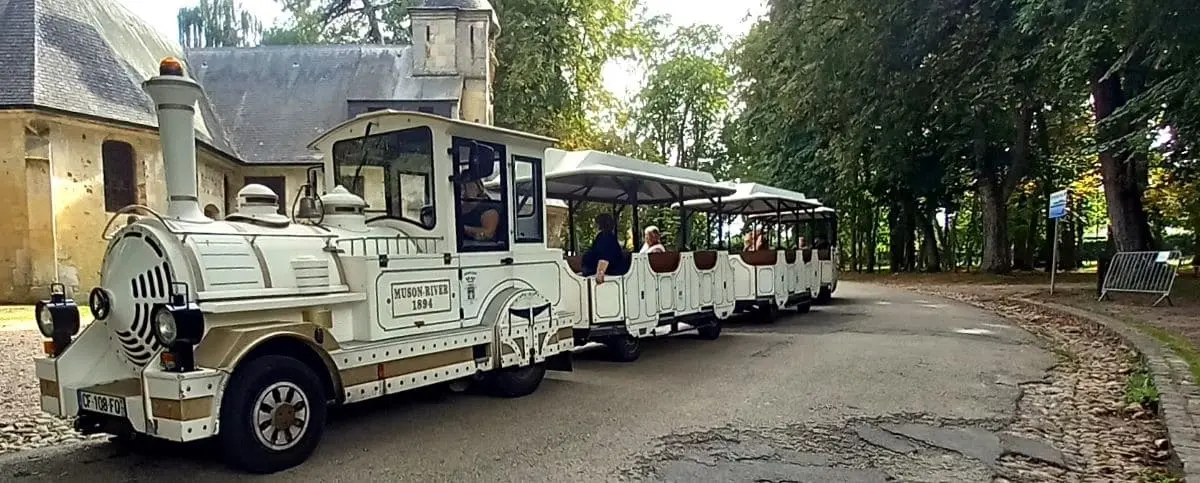 A white touristic train with multiple open carriages, filled with passengers. The train is driving through a scenic area with trees and historical buildings.