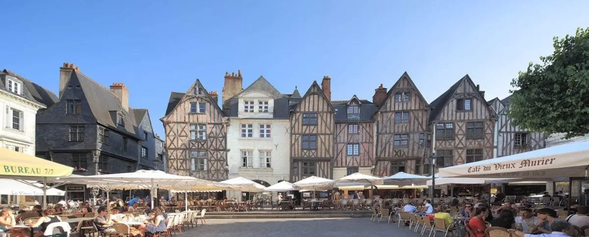 A bustling city center in Tours, France, with people dining at outdoor cafes, surrounded by charming, half-timbered medieval buildings under a clear blue sky.
