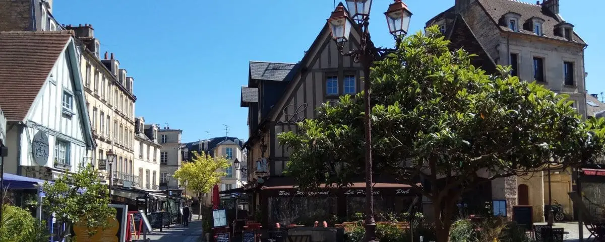 A charming town center in Caen with historic buildings, outdoor cafes, and leafy trees. The clear sky and people walking around create a welcoming atmosphere.