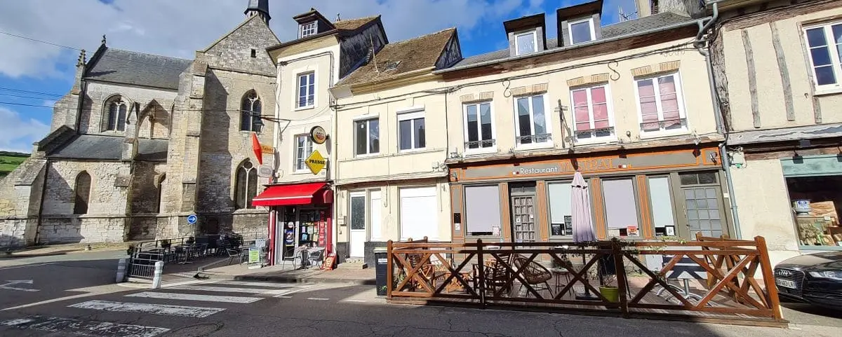 A vibrant view of the town center in Les Andelys, featuring historic buildings, a church, a small outdoor seating area, and shops along a quiet street.