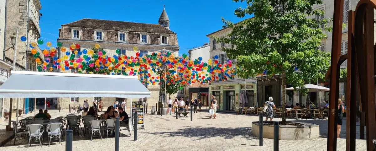 A lively town square in Périgueux, adorned with colorful hanging balloons and outdoor seating. People are enjoying the sunny day in the pedestrian area, surrounded by historic buildings.