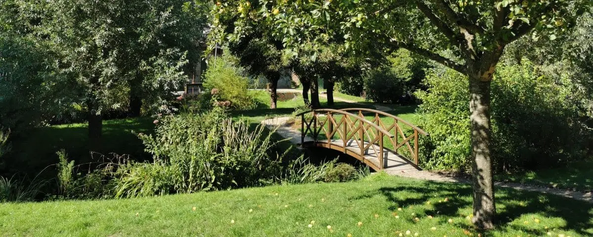 A scenic park with a small wooden bridge over a stream, surrounded by lush greenery, trees, and a well-kept lawn, creating a peaceful and inviting setting.