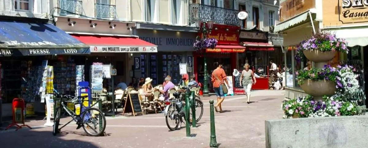 A lively scene in the town center of Trouville, with people walking, cafes with outdoor seating, bicycles, and vibrant storefronts under sunny weather.
