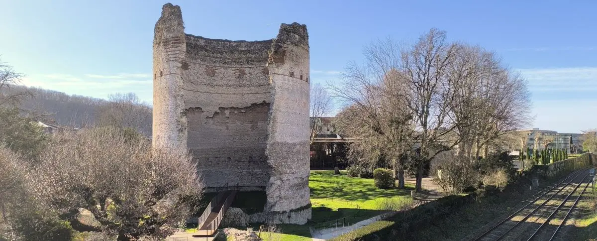 The remains of Vesone Tower in Périgueux, featuring a large, circular stone structure with weathered surfaces. The tower stands amidst a landscaped area with a pathway and greenery.
