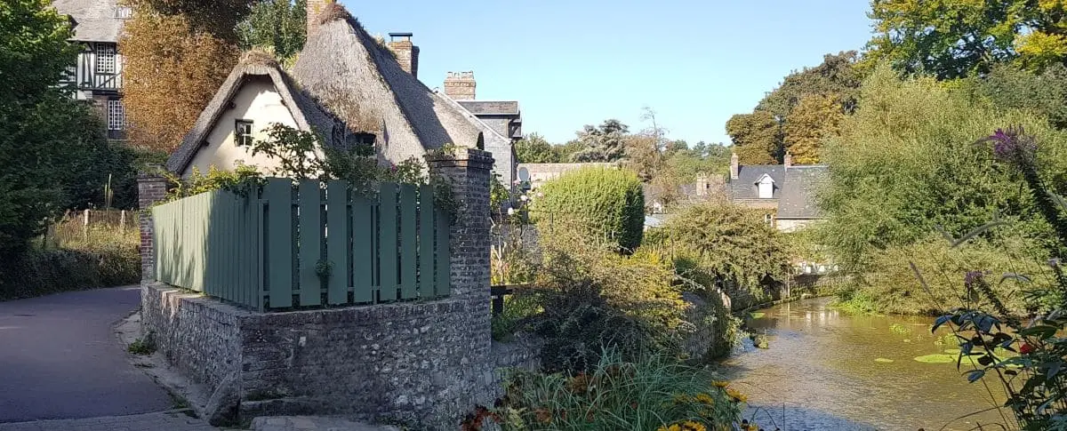 Charming cottage with a thatched roof in Veules-les-Roses village, nestled along a quiet river and surrounded by greenery and colorful flowers.