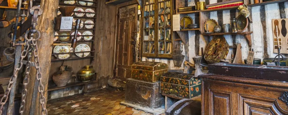 An interior view of the Vieux Honfleur Museum, displaying historical artifacts including plates, scales, and chests. The room has a rustic feel with wooden beams and a variety of old objects.