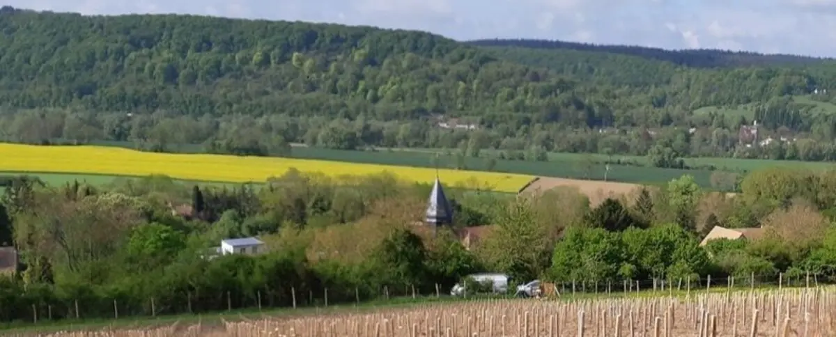 A scenic view of the countryside in Giverny, featuring rolling hills, patches of farmland, and a small village with a church steeple in the distance. The landscape is lush and green, with fields of yellow flowers visible.