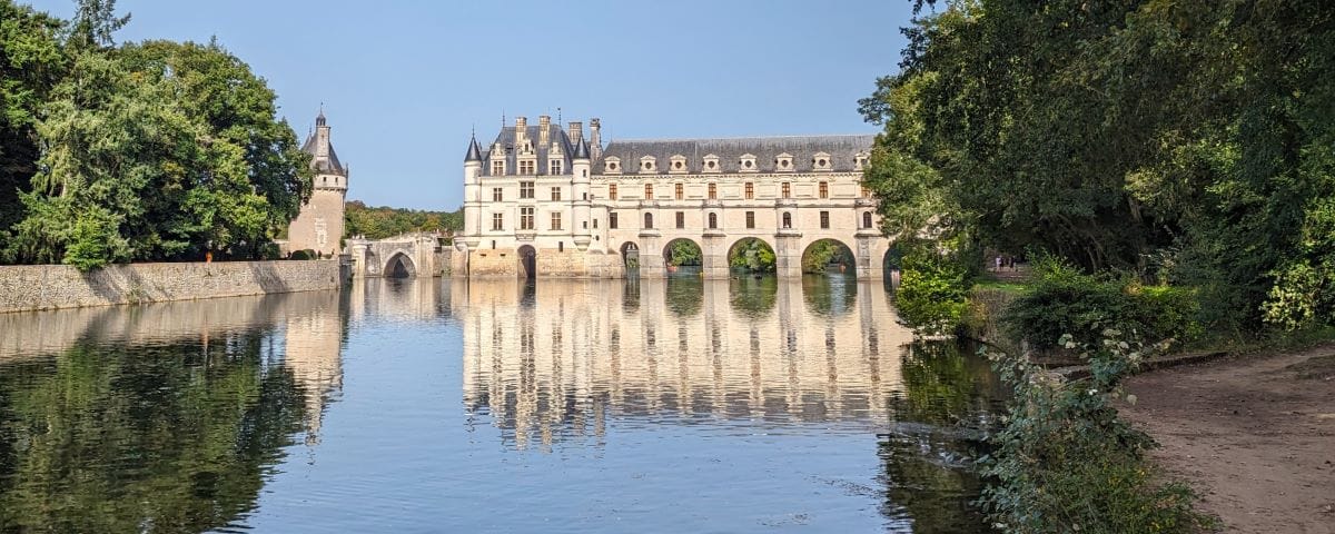 A scenic view of the Château de Chenonceau spanning the Cher River, with lush greenery on both sides. The castle's arches are reflected in the still waters, creating a picturesque scene.