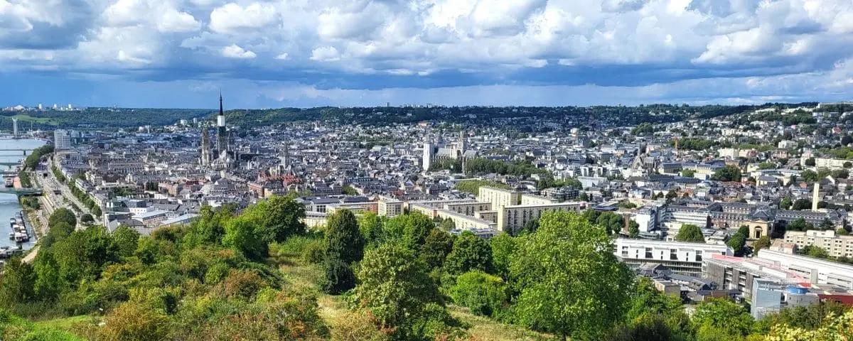 A panoramic view of Rouen from the Côte Sainte-Catherine viewpoint, showing the cityscape, including the Rouen Cathedral and the winding Seine River, under a dramatic sky with scattered clouds.