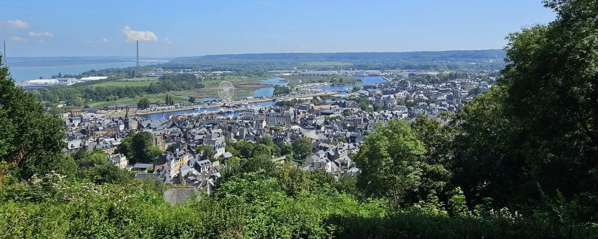 An elevated view from Mont Joli viewpoint, overlooking the town of Honfleur with its clustered buildings, winding streets, and surrounding countryside. The scene is framed by green trees.