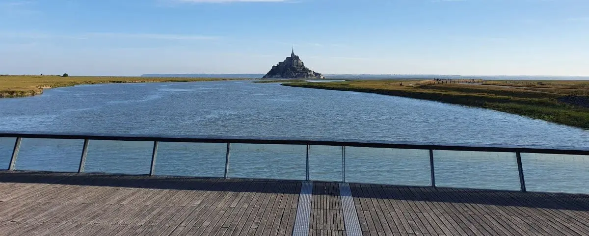 A distant view of Mont Saint-Michel from a wooden deck, with the abbey rising above the surrounding water and grasslands.