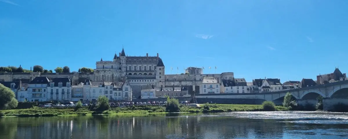 A scenic view of Amboise Castle from across a river, with the grand castle and its surrounding buildings reflecting in the calm water. The sky is clear, enhancing the castle's majestic presence.
