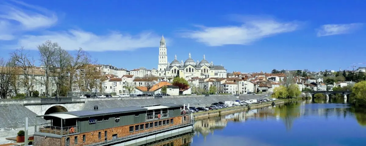 A panoramic view of Périgueux from Saint-Georges Bridge, with the Périgueux Cathedral prominently visible against the skyline. The scene captures the river, waterfront buildings, and a clear blue sky.