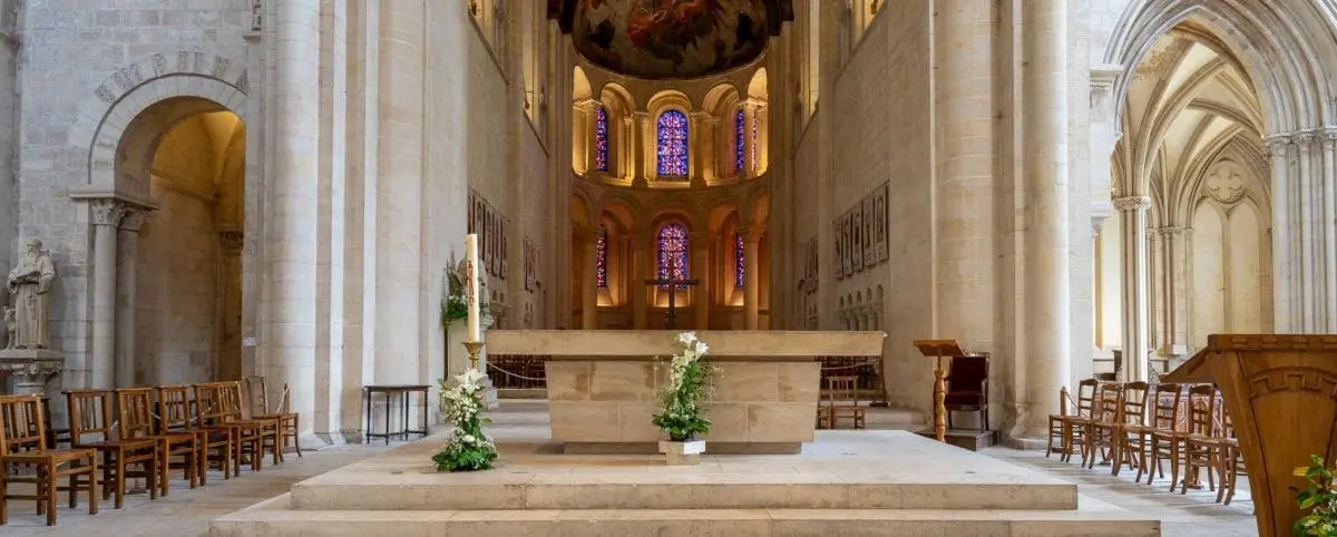 The interior of the Women's Abbey in Caen, showing its grand altar and gothic architecture with high arches and stained glass windows. The arrangement of chairs and flowers adds to the solemnity of the space.